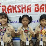 Three young girls in beige school uniforms pose with decorative medals in front of a large banner wearing id lanyards and smiling lightly